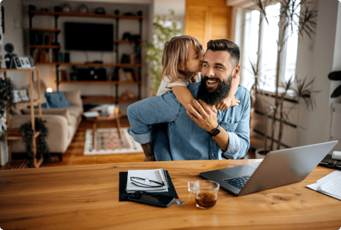 Man sitting at kitchen table working on laptop with his daughter hugging him